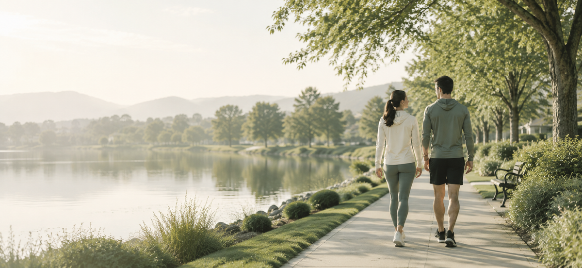 A man and a woman walk away on a paved path beside a calm lake, with trees, hills, and a park bench in the background.