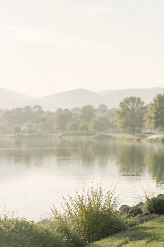 A serene outdoor landscape with a calm lake, distant tree-lined shore, faint mountains, and soft misty light over the scene.