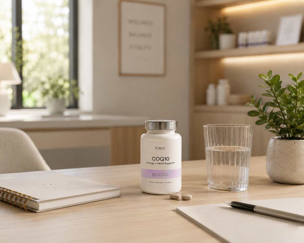 A white bottle of PURA COQ10 dietary supplement with a silver cap and lavender label, placed on a light wood desk next to a glass of water, with a notebook and pen in the foreground and a blurred home office background.